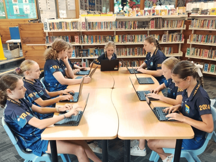 Eight students working on laptops in the library with shelves of books in the background,