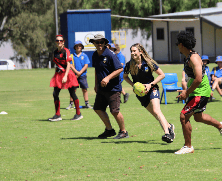 Teachers and students playing football on a large oval.