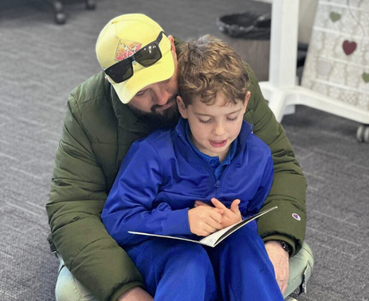 A student sitting on their father's lap and reading a book.
